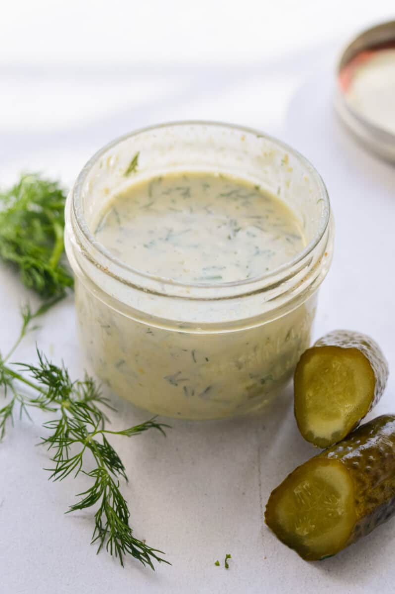Jar of dill pickle salad dressing next to sprigs of dill and a pickle.