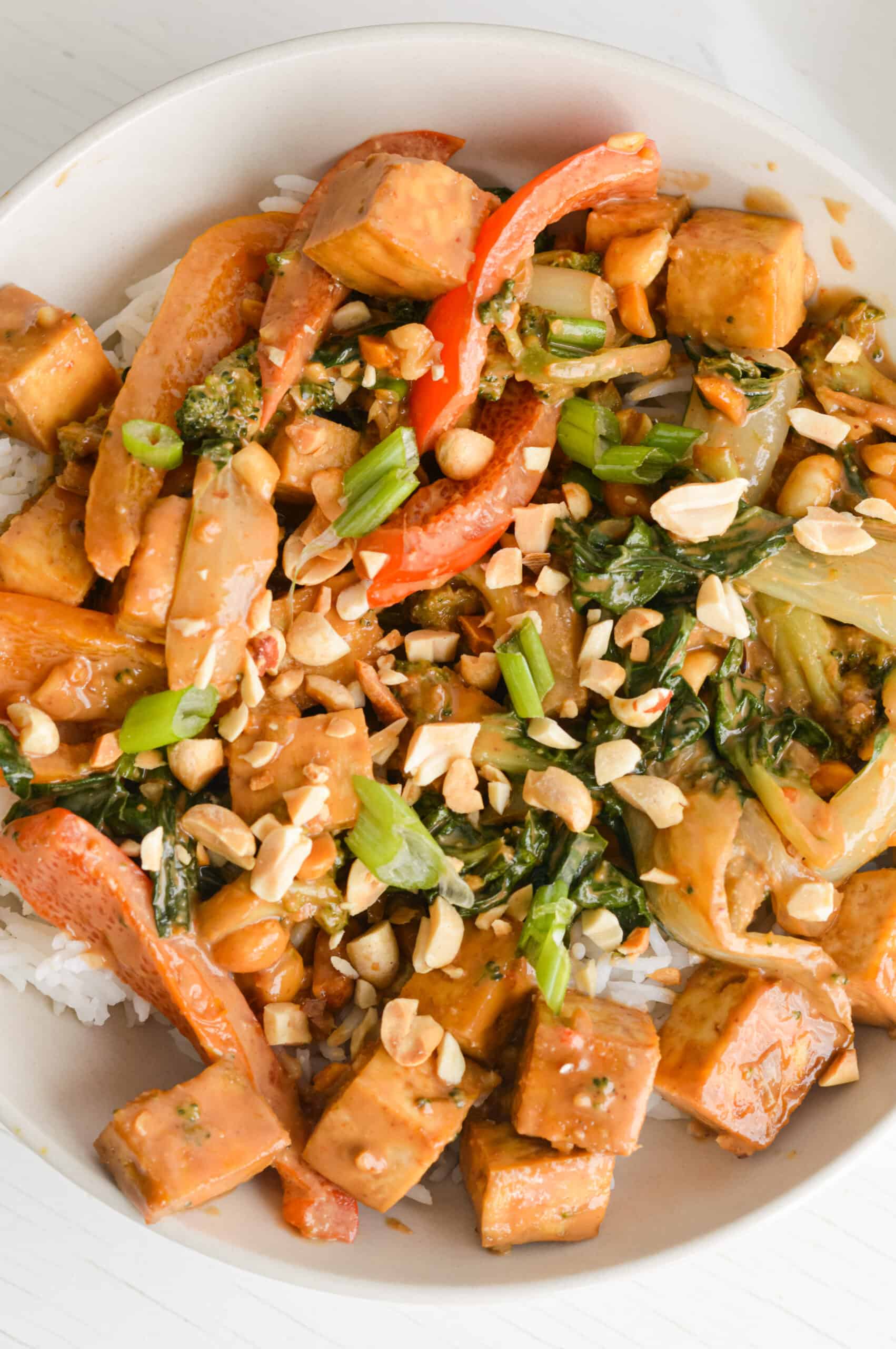Bowl of tofu satay stir fry with bowls of rice and crushed peanuts in the background.