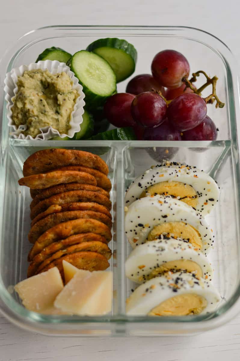 Adult lunchable in a glass container with boiled eggs, crackers, cheese, grapes, cucumbers and dip.