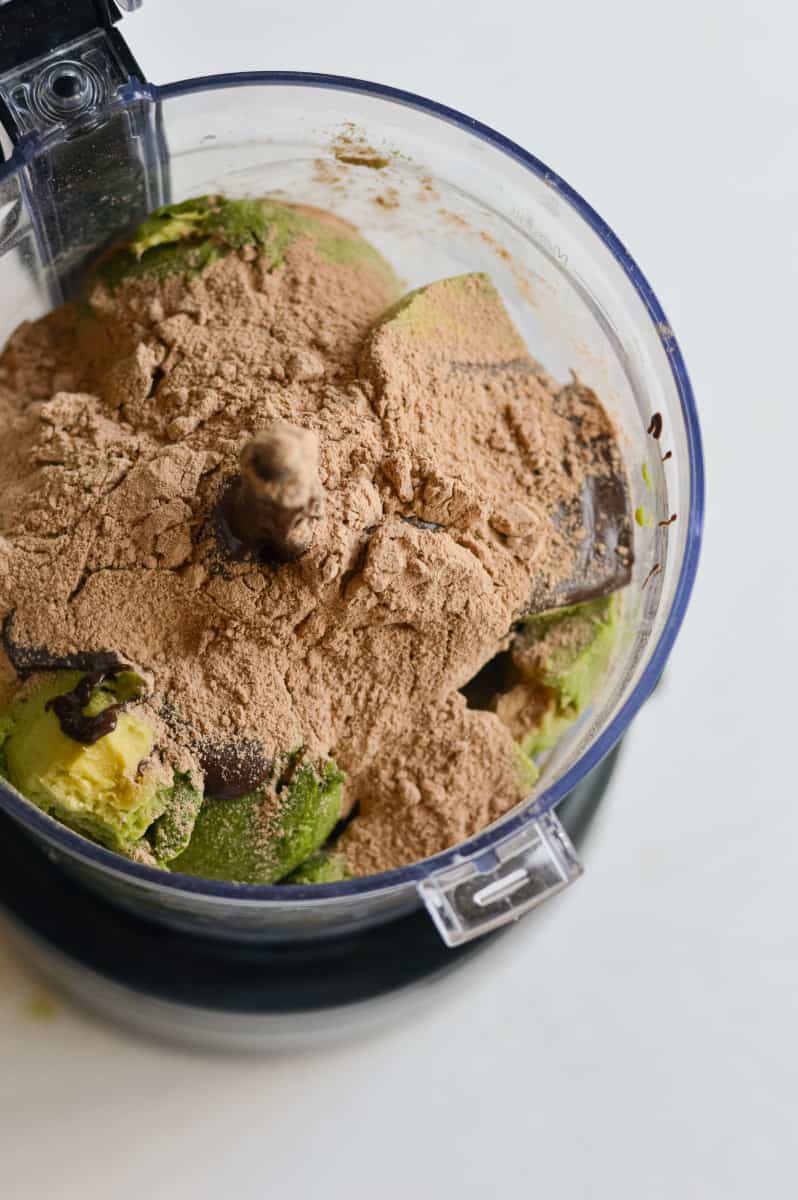 Adding chocolate protein powder to avocados in a food processor.