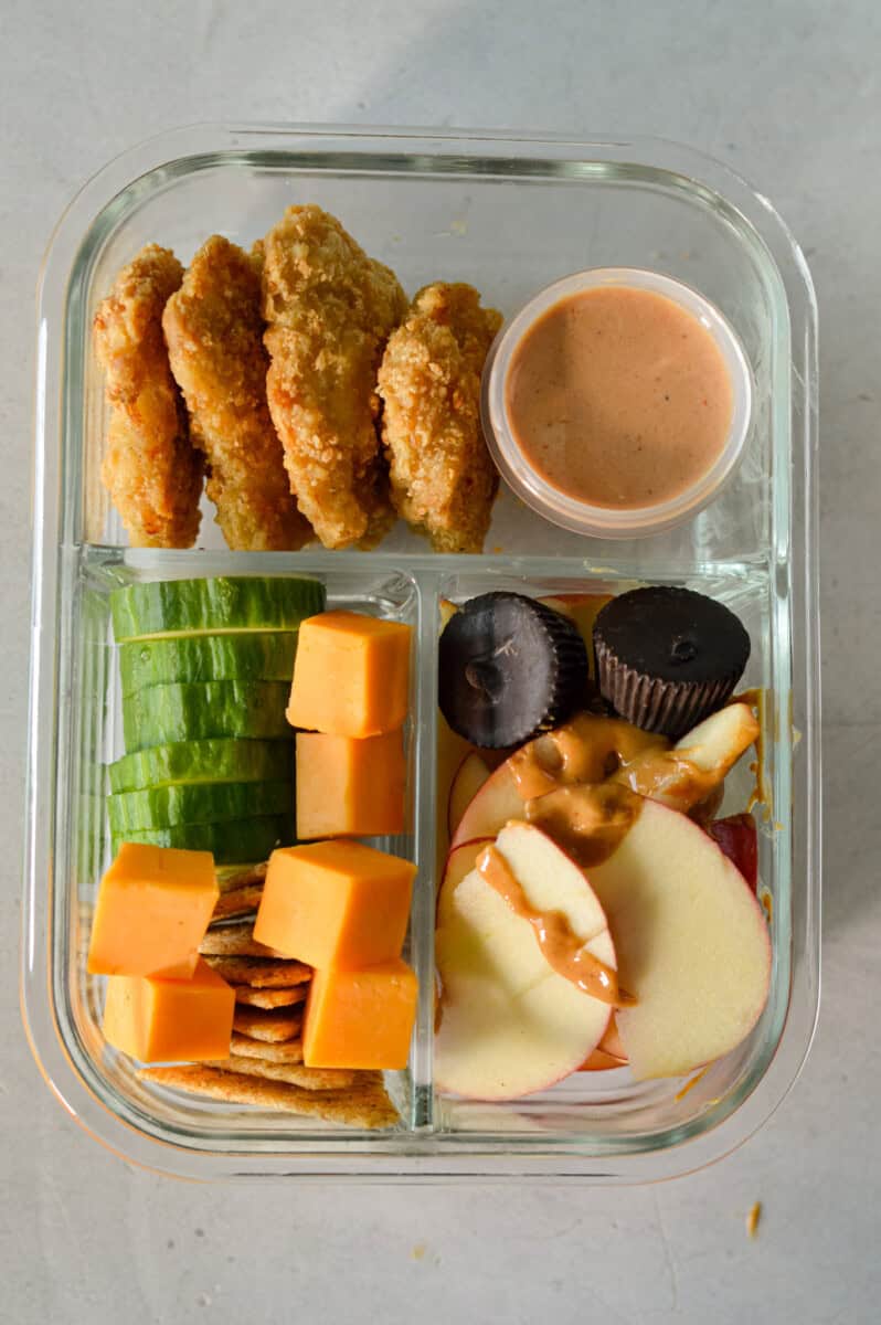 Adult lunchable with chicken nuggets, dip, cheese, crackers, cucumbers, chocolate, apples and peanut butter.