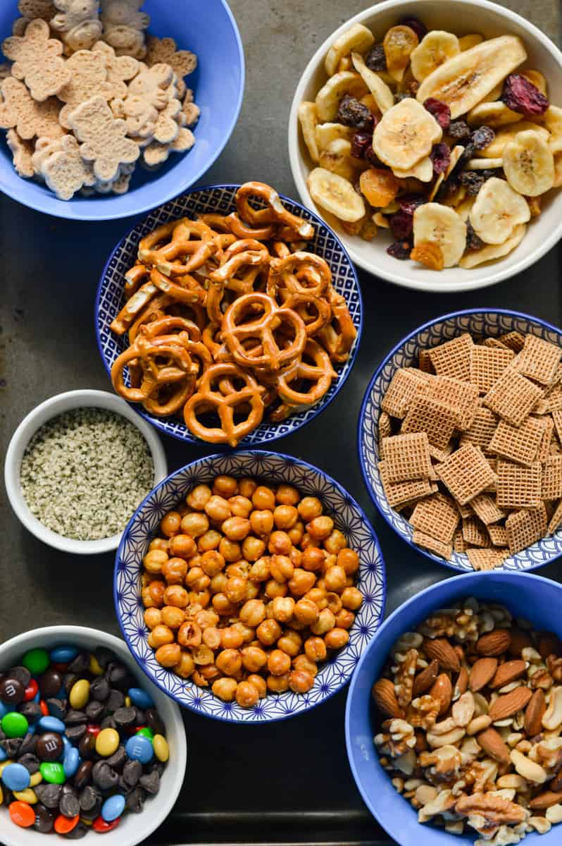 Ingredients for trail mix including popcorn, pretzels, chocolate, cereal, hemp seeds and dried banana.