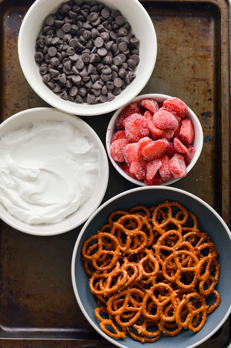 Ingredients in bowls, including chocolate chips, yogurt, strawberries and pretzels.
