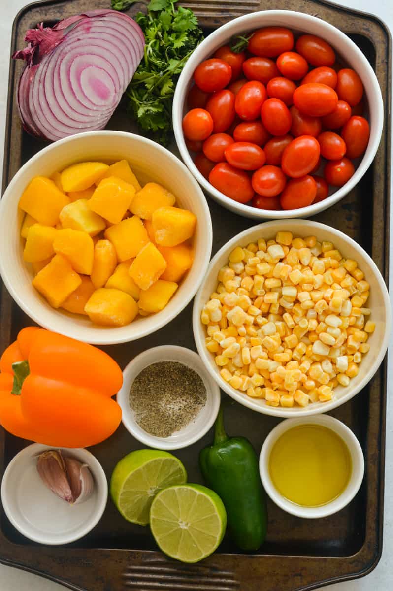 Ingredients including cherry tomatoes, corn, red onion, pepper, jalapeno, mango, seasoning and lime.