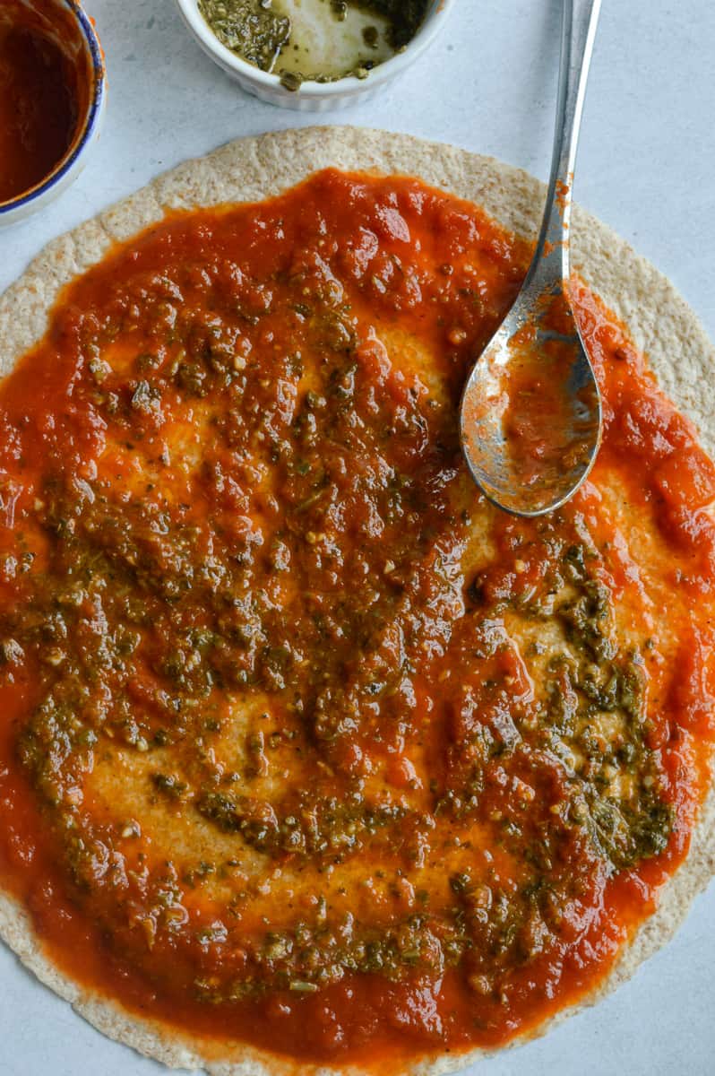 Spreading tomato sauce and pesto on  whole wheat tortilla.