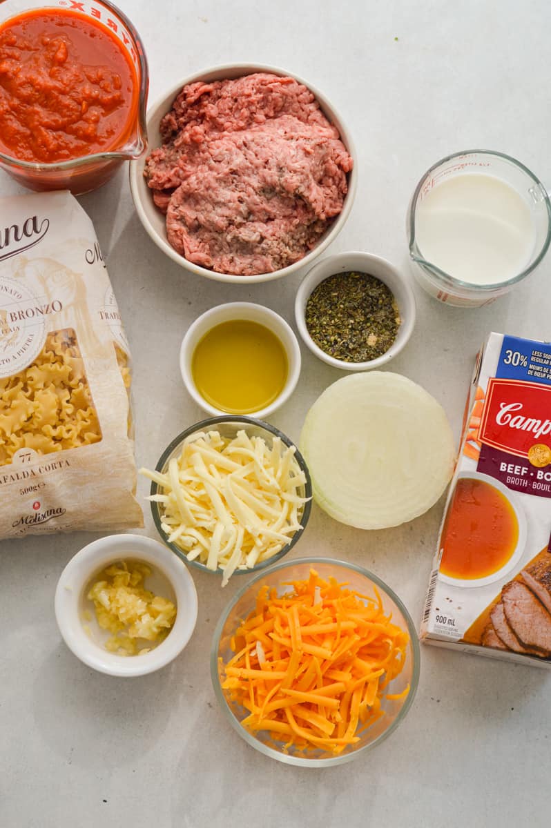 Ingredients including mafalda pasta, ground beef, cheeses, seasoning, garlic, olive oil, spinach, onions. shredded veggies, broth and milk.