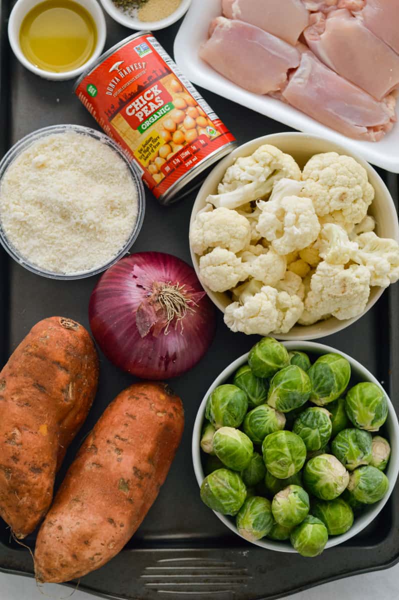 Ingredients including chicken thighs, red onion, brussel sprouts, sweet potato, cauliflower, parmesan, seasoning and olive oil.
