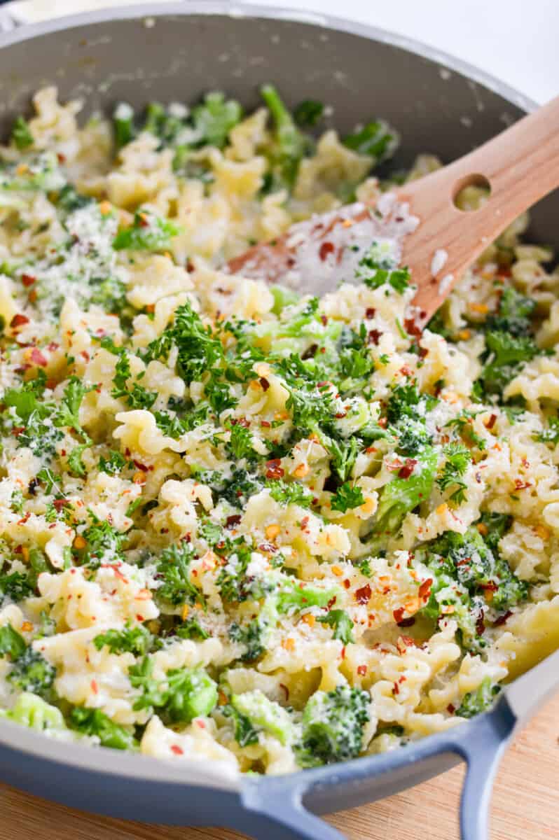 Adding broccoli and pasta to alfredo sauce, garnishing with parsley and red pepper flakes.