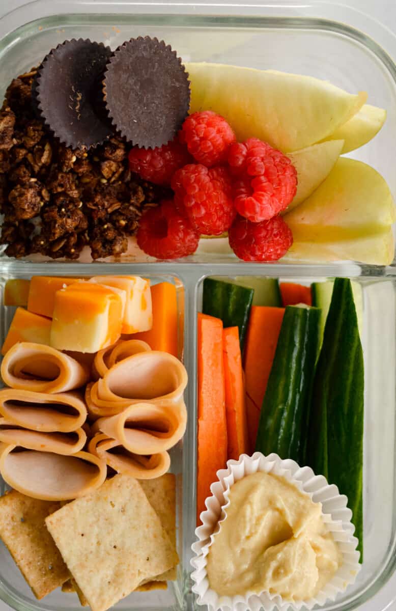 Adult lunchable in glass container with deli meat, carrots, hummus, fruit, cheese, cucumber, chocolate and granola.