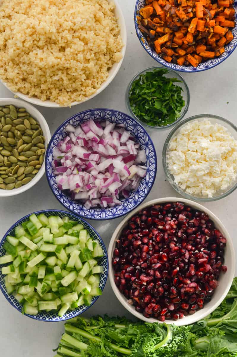 Ingredients including kale, pomegranate, cucumber, red onion, parsley, feta, pumpkin seeds, quinoa, and sweet potato.