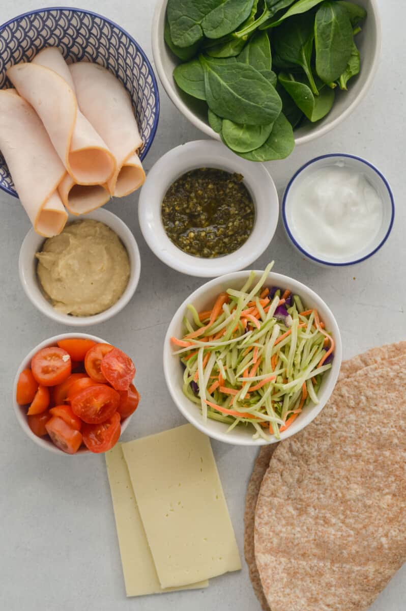 Ingredients including coleslaw, deli chicken breast, hummus, Greek yogurt, seasoning, cherry tomatoes, cheese, tortillas, pesto and spinach.
