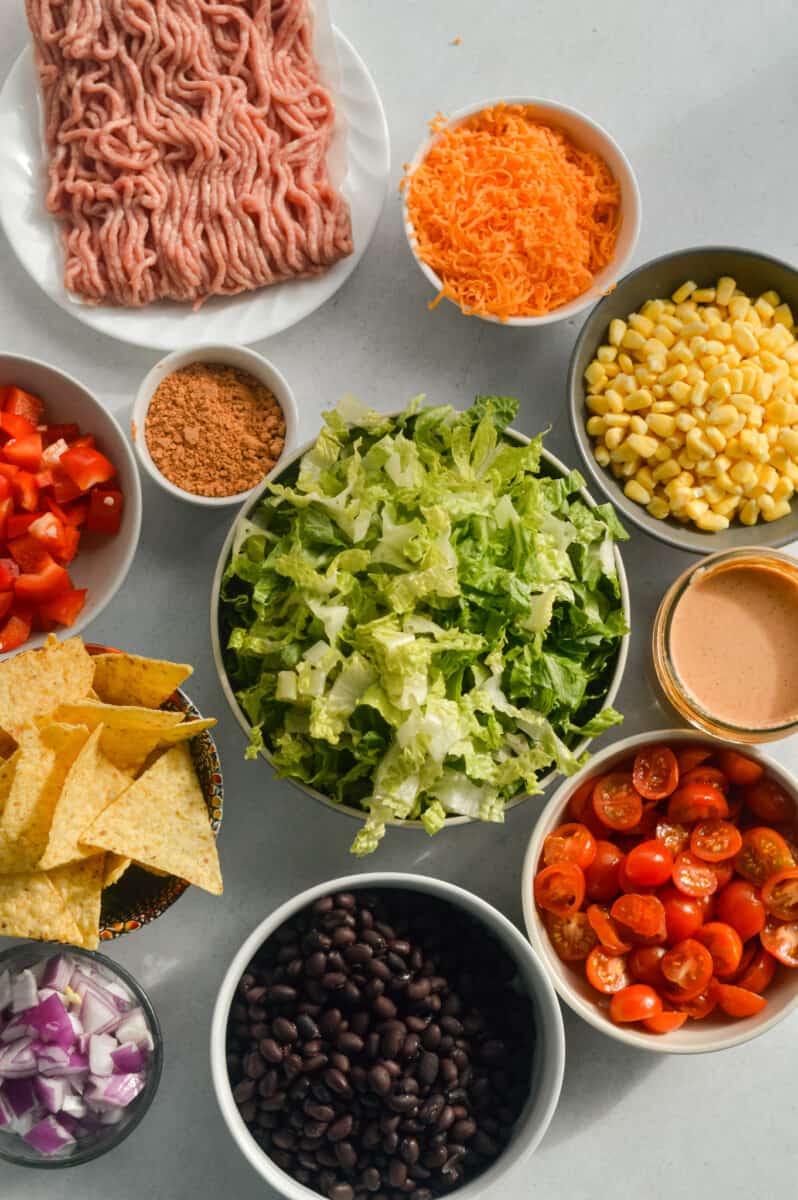 Ingredients including ground turkey, romaine lettuce, corn, cherry tomatoes, bell pepper, cheese, seasoning, black beans and salsa dressing.