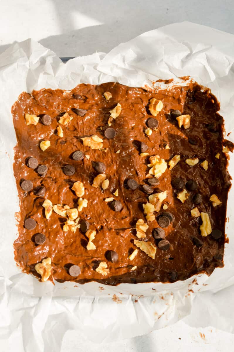 Brownie batter in a parchment lined tray, topped with walnuts and chocolate chips.