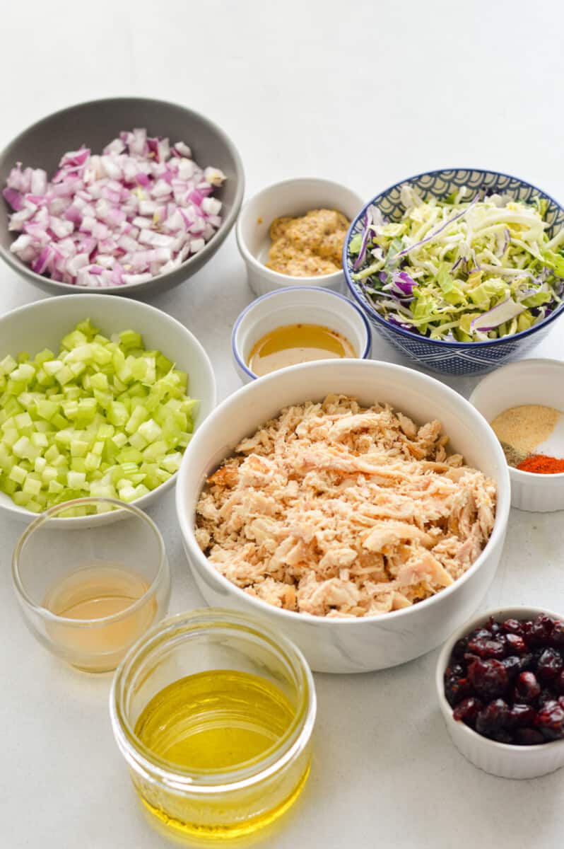 Ingredients including shredded chicken, diced celery, diced red onion, kale slaw, olive oil, honey, dijon mustard, spices and cranberries.