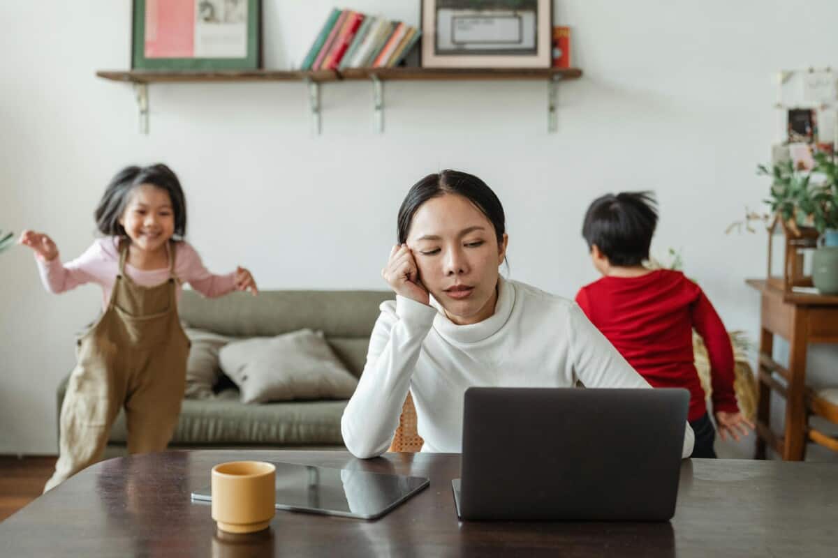 Stressed mom staring at a laptop with kids running behind her.