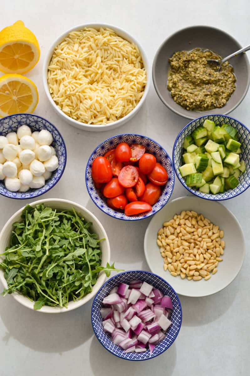 Measured ingredients including bocconcini, pesto, orzo, cherry tomatoes, cucumbers, red onion, pine nuts, arugula, and lemon.