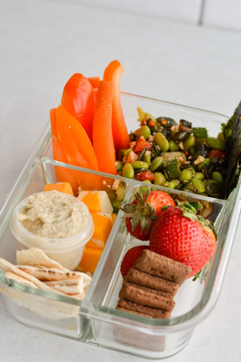Vegetarian bento box with edamame salad, veggies, fruit, cheese, pita and hummus.