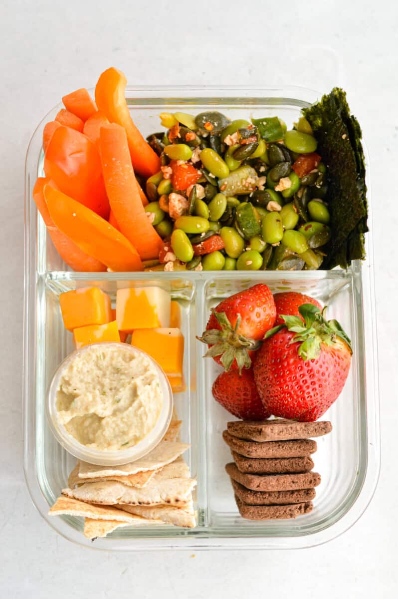 Birds eye of vegetarian bento box with carrots, strawberries, cheese, hummus, pita, cookies and edamame salad.