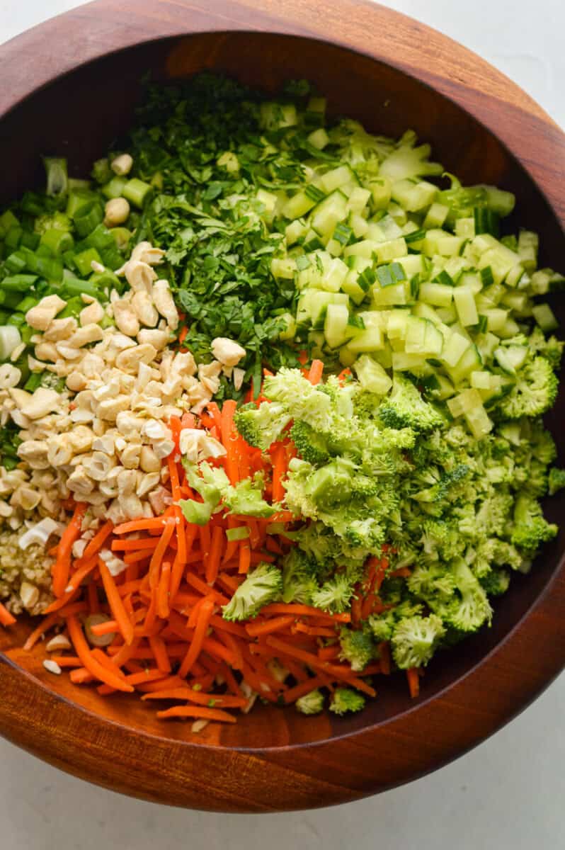 Adding cooked quinoa, shredded cabbage, cilantro, shredded carrots, broccoli, bell peppers and green onions to a bowl.

