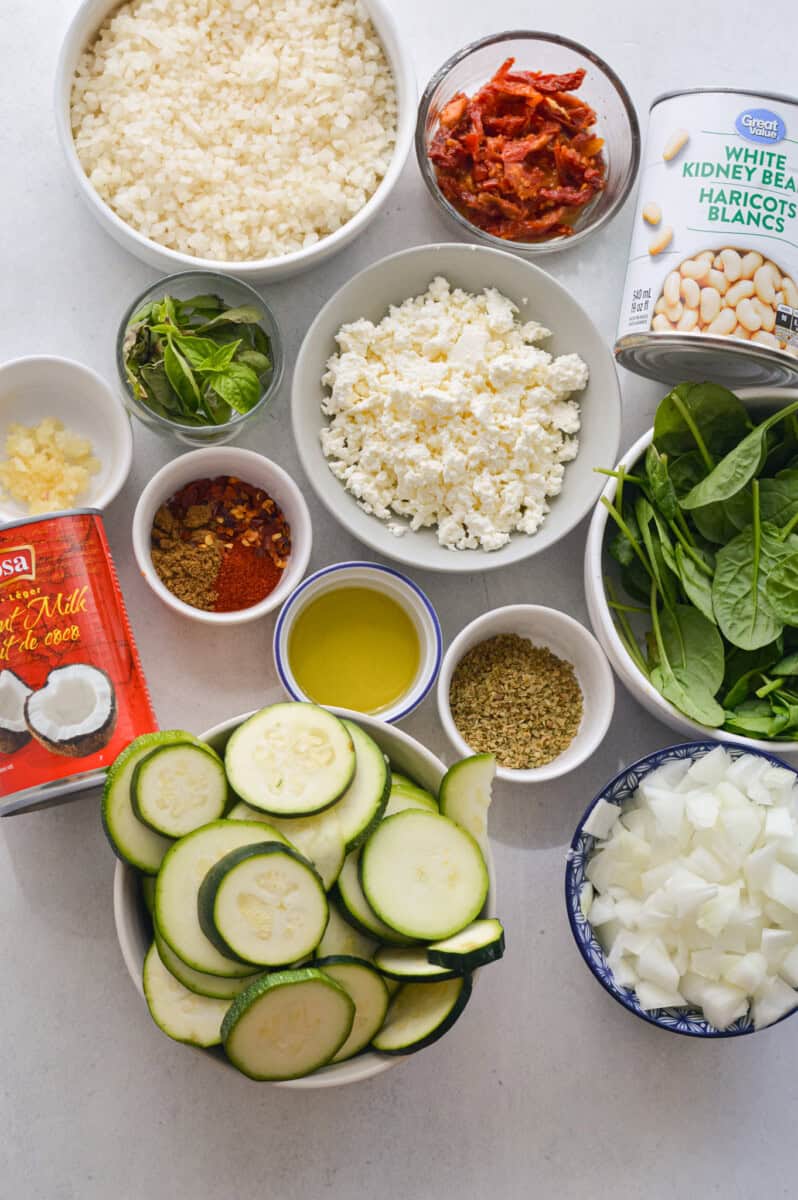 Ingredients including olive oil, zucchini, onion, cauliflower rice, coconut milk, feta cheese, spices, basil, spinach, white beans, sundried tomato paste and garlic.