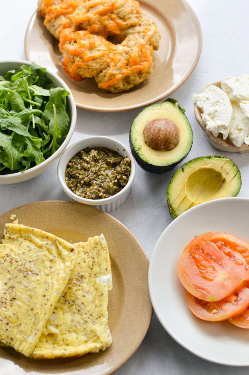 Ingredients including avocado, tomato, bagel, arugula, feta, pesto and eggs.