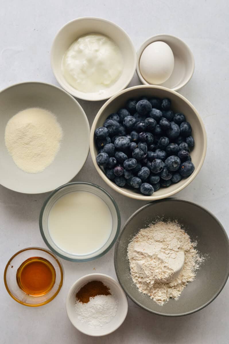 Ingredients including blueberries, strawberries, banana slices, flour, protein powder, greek yogurt, milk, maple syrup, baking powder and cinnamon.