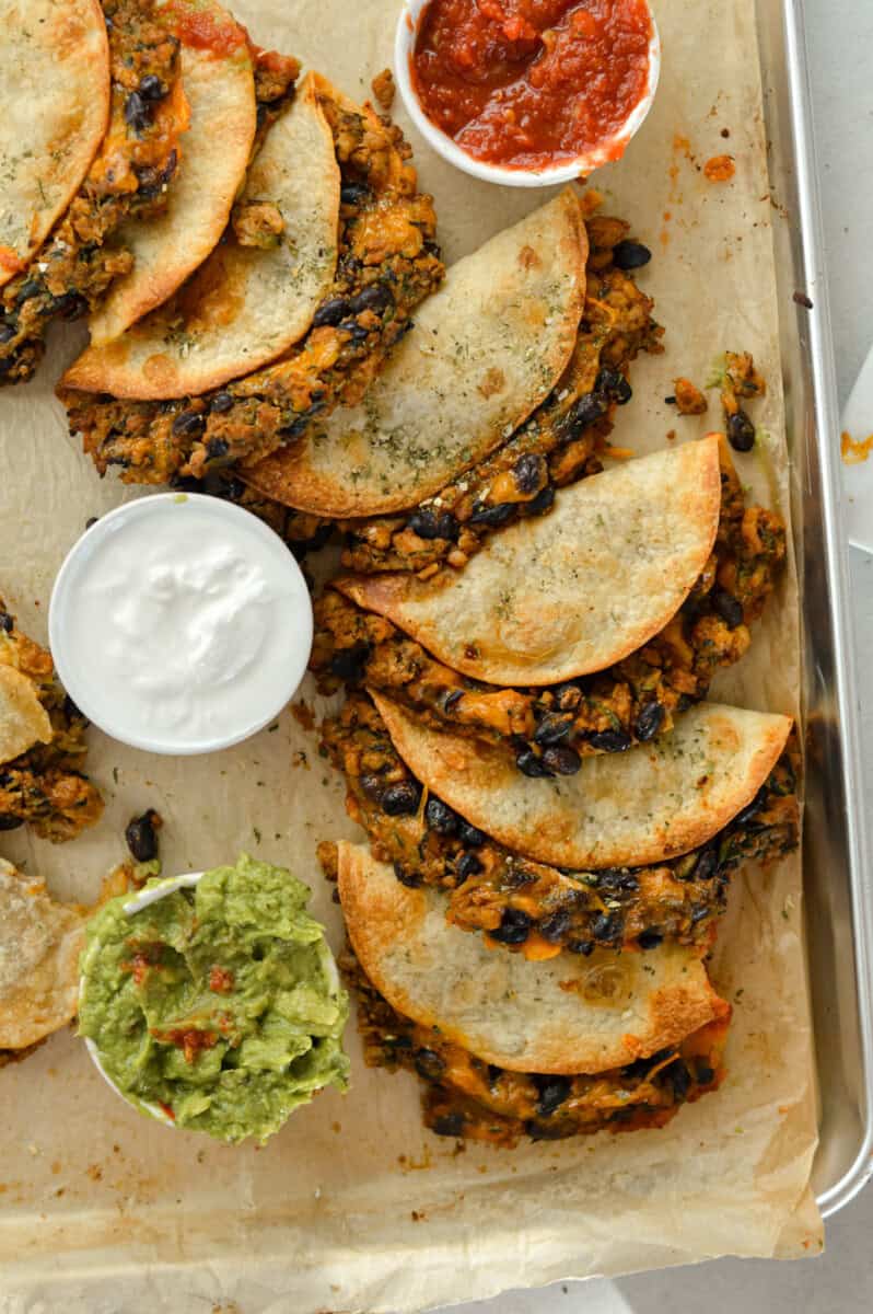 Birds eye of crispy baked chicken tacos on a sheet pan, served with salsa, guacamole and sour cream.