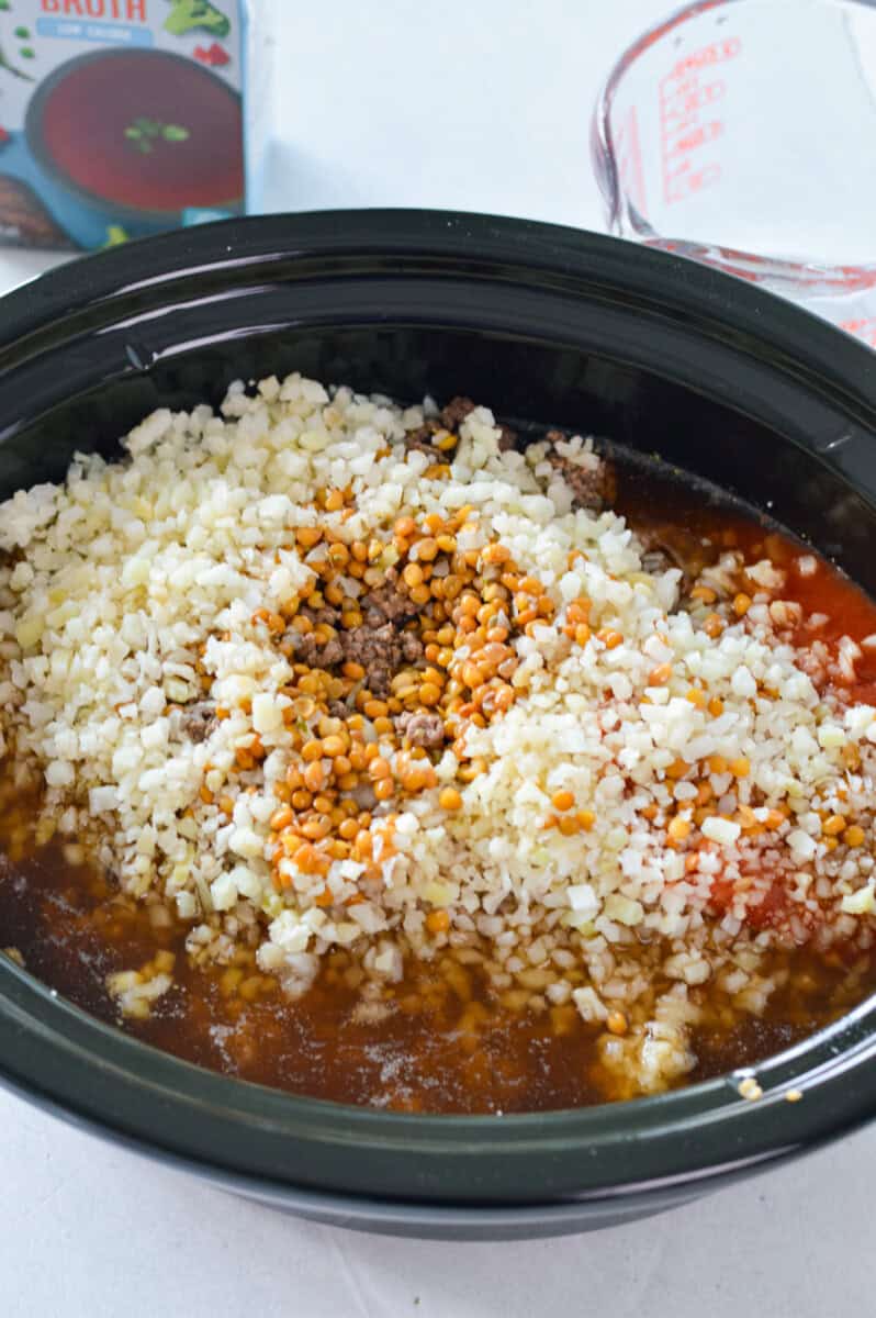 Adding cooked ground beef, diced yellow onion, green pepper, red pepper, diced tomatoes (and their juices), beef broth, tomato sauce, oregano, garlic powder, salt and pepper (to taste), worcestershire, riced cauliflower and lentils to a slow cooker.