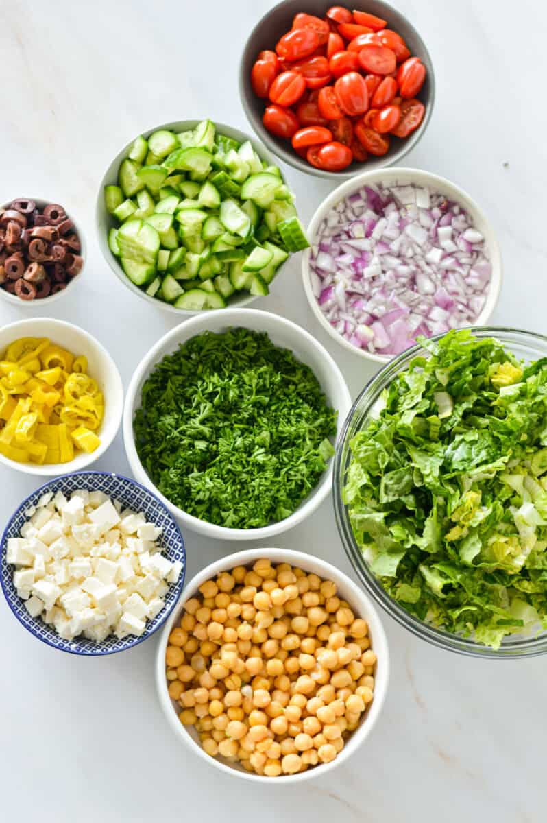 Ingredients including chopped romaine lettuce, red onion, cucumber, chickpeas, cherry tomatoes, pepperocinis, feta cheese and olives.