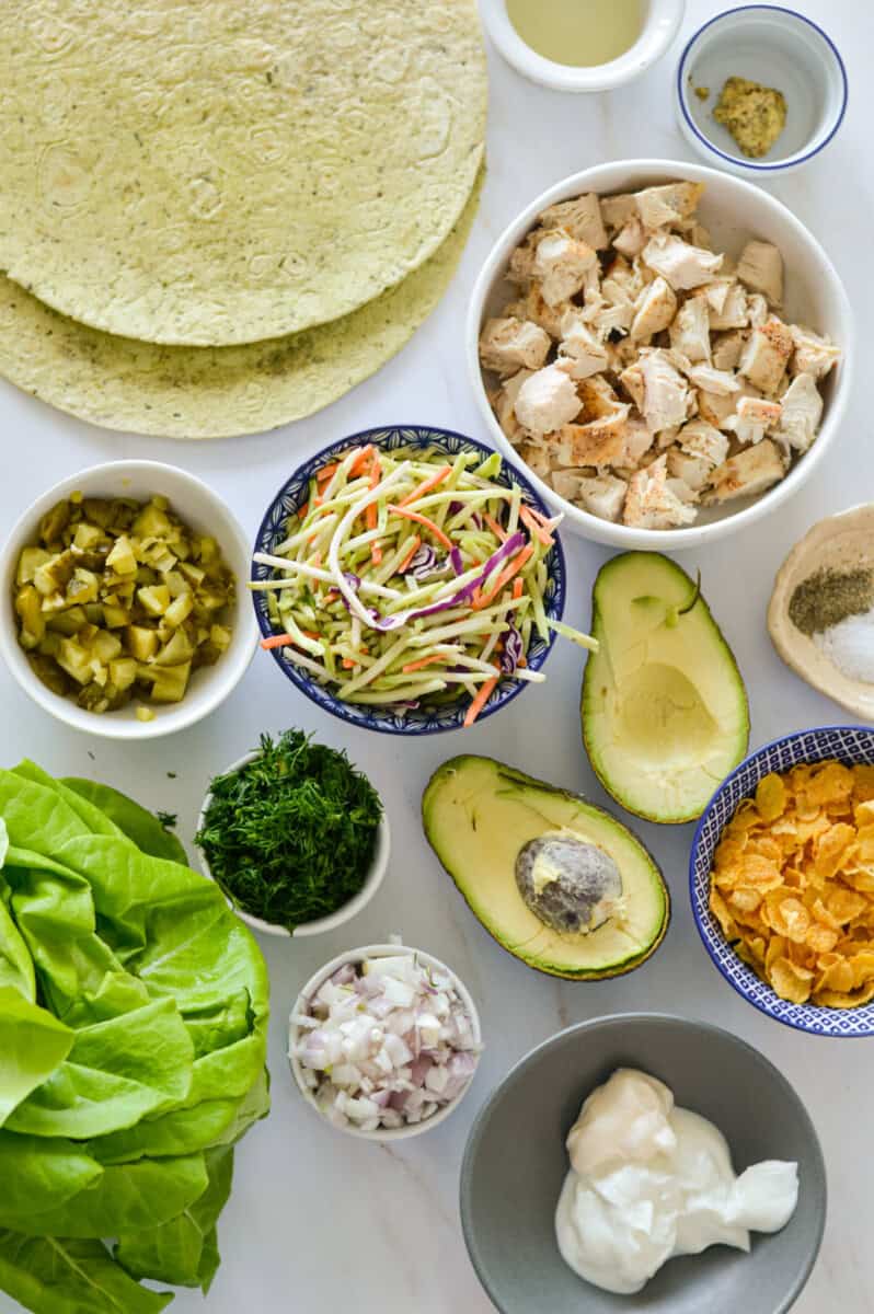Ingredients including chicken, avocado, dill, pickles, coleslaw, tortillas, shallot, mustard, spices, greek yogurt, mayonnaise and lettuce.
