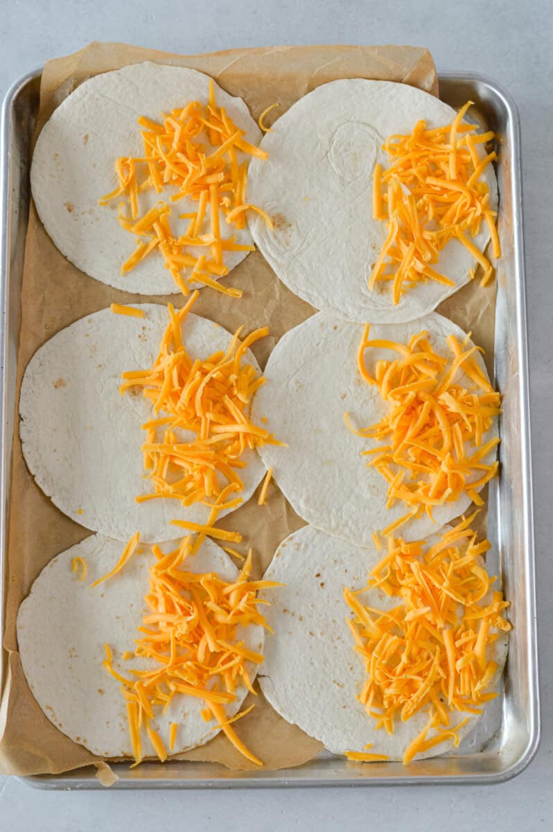 Addnig shredded cheese to 6 small tortillas on a tray.