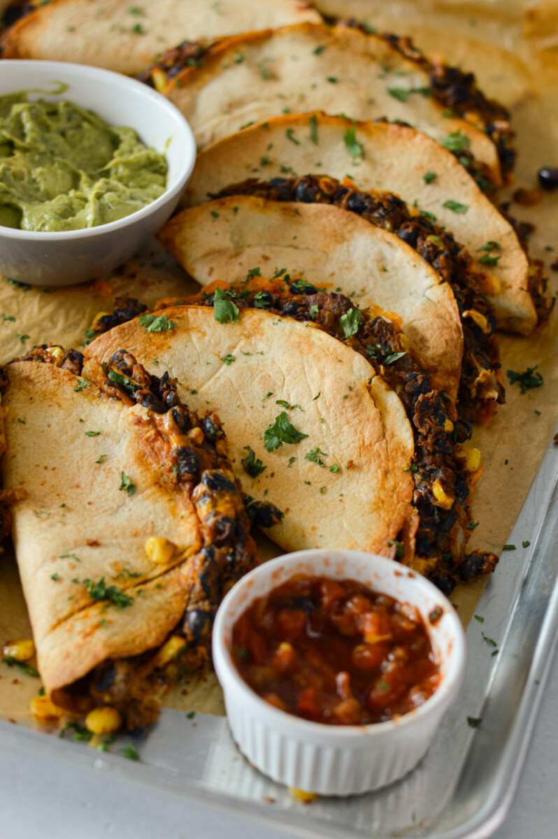 Close up of black bean tacos on a sheet pan with guac and salsa.