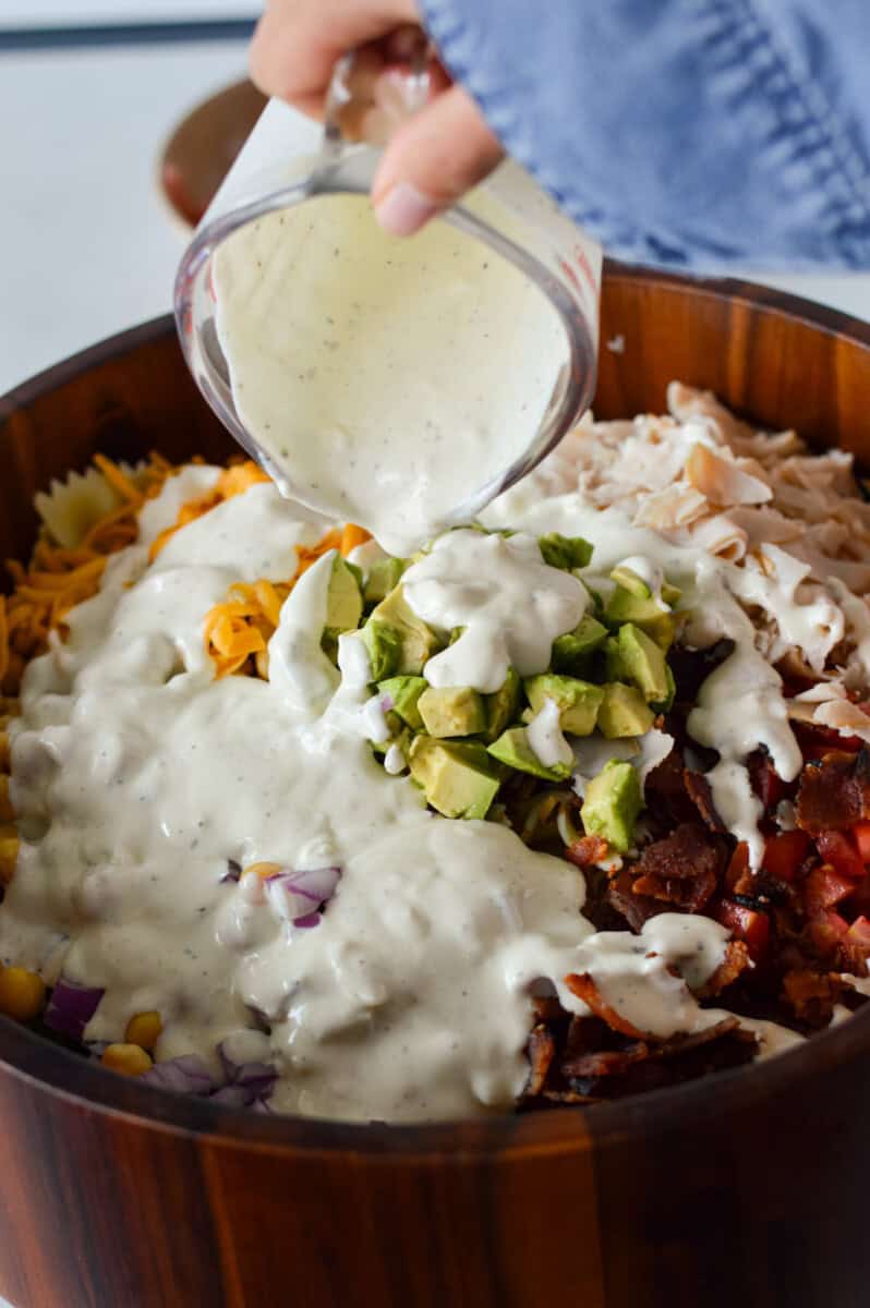 Pouring greek yogurt on a bowl of BLT pasta salad.
