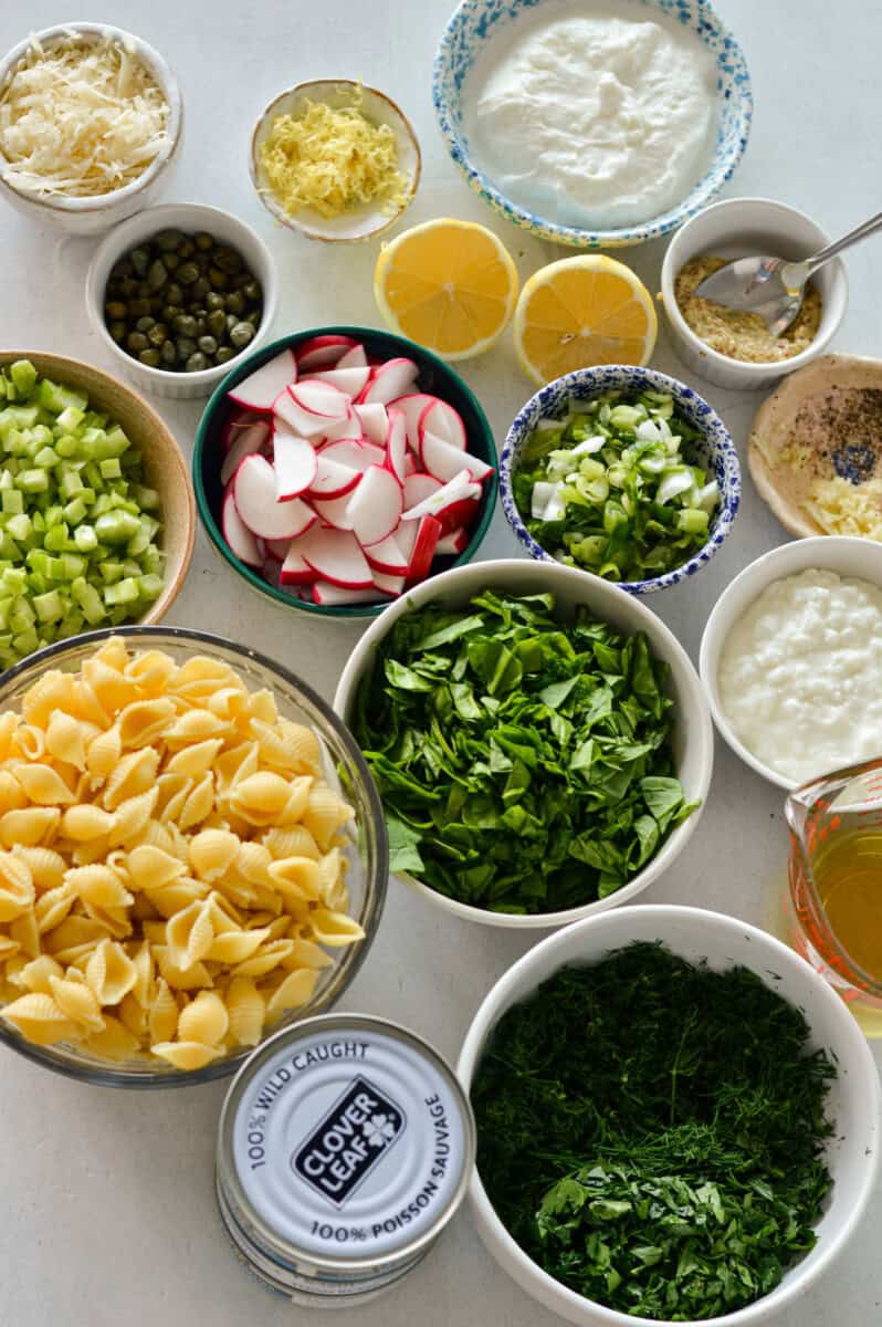 Ingredients including pasta, canned tuna, celery, spinach, radishes, dressing, capers and parmesan.