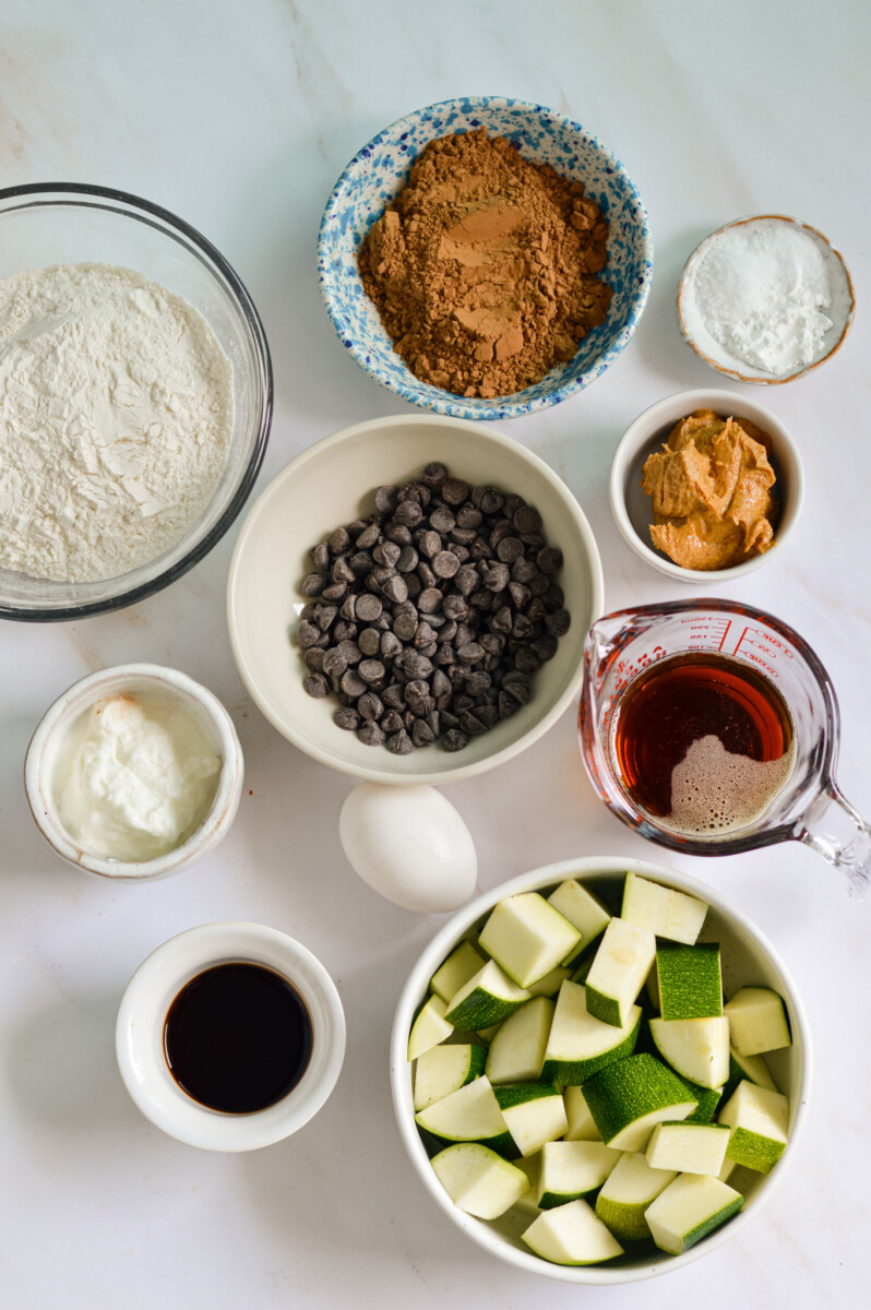 Ingredients including flour, cocoa powder, zucchini, maple syrup, greek yogurt, peanut butter, eggs, chocolate chips.