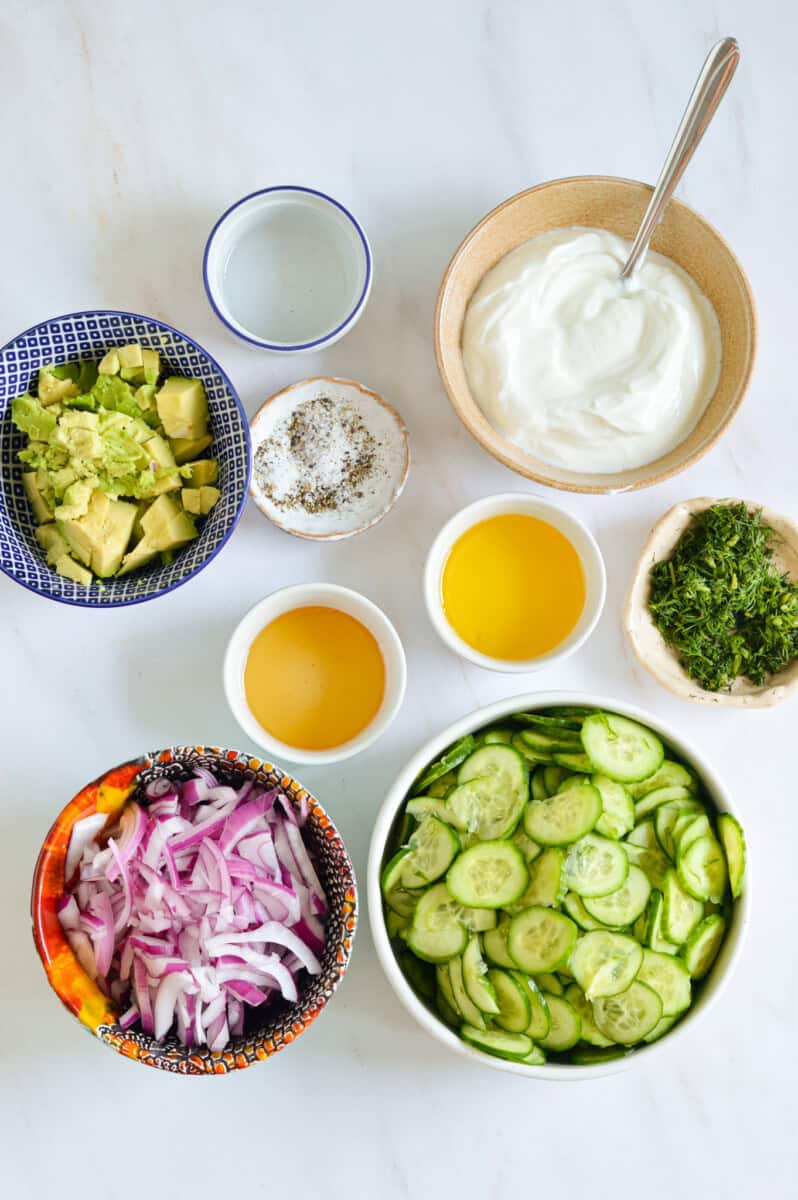Ingredients including sliced cucumber, red onion, avocado, fresh herb, olive oil, lemon juice, salt and pepper and greek yogurt.
